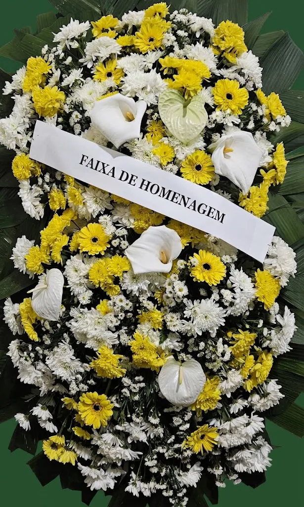 Coroa de gérberas amarelas com copos-de-leite, antúrios brancos e faixa de homenagem